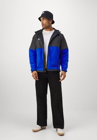 Sort og blå tofarvet jakke med lynlås og hætte samt Nike-logo, båret med sorte bukser og hvide sneakers. Sort bøttehat.