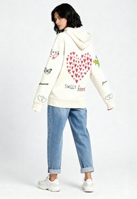 Kremasti hoodie s crvenim srcem, skicama i tekstom na stražnjoj strani; mekani materijal, širok kroj, u kombinaciji s lagano podvrnutim svijetloplavim trapericama i tenisicama.