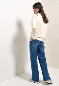 Beige top met lange mouwen en een ontspannen pasvorm, gecombineerd met losse blauwe jeans en grijze sneakers. Het haar is halflang en bruin.