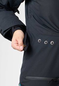 Zwarte waterdichte jas met een hand die een pocket vastgrijpt. Voorzien van drukknoopsluitingen en een ritszak aan de zijkant voor veilige opslag.
