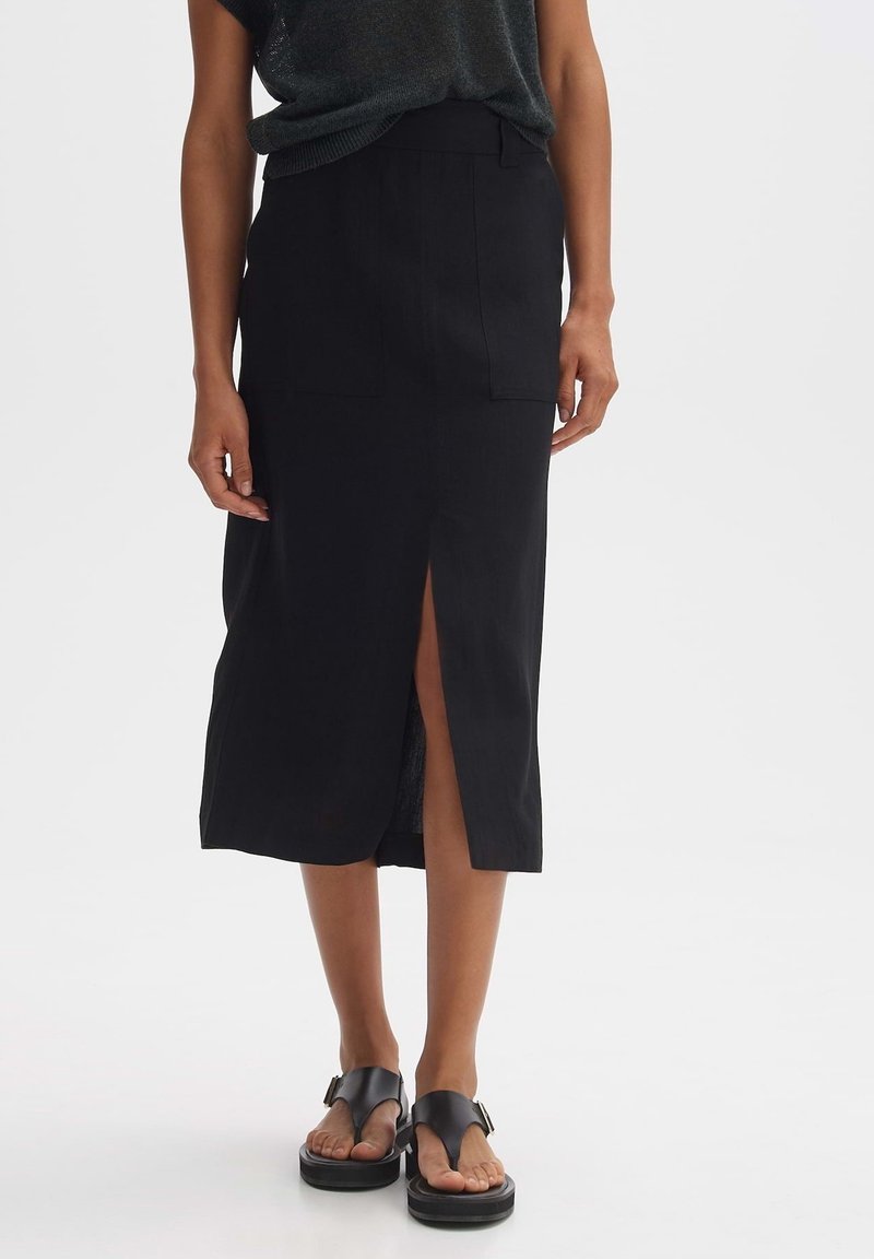 Femme portant une jupe midi noire avec une fente sur le devant, associée à des sandales compensées noires, debout devant un fond blanc.