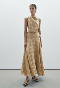 Langes beiges Kleid mit einem strukturierten, gewebten Muster, rundem Ausschnitt und kurzen Ärmeln. Kombiniert mit schwarzen Riemchensandalen.