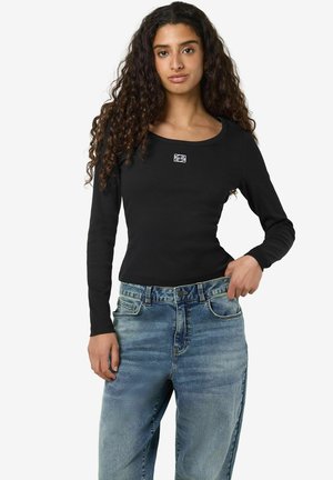 Črn dolgosleevni top z okroglo ovratnico in manjšim logotipom, v kombinaciji z svetlomodrimi izblukanim jeansom z višjim pasom in klasičnim petimi žepi.