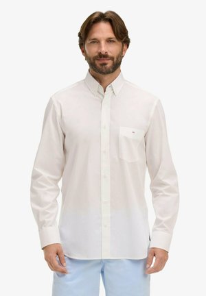 Homme portant une chemise blanche à manches longues boutonnée avec une poche poitrine et un pantalon bleu clair, debout devant un fond blanc.