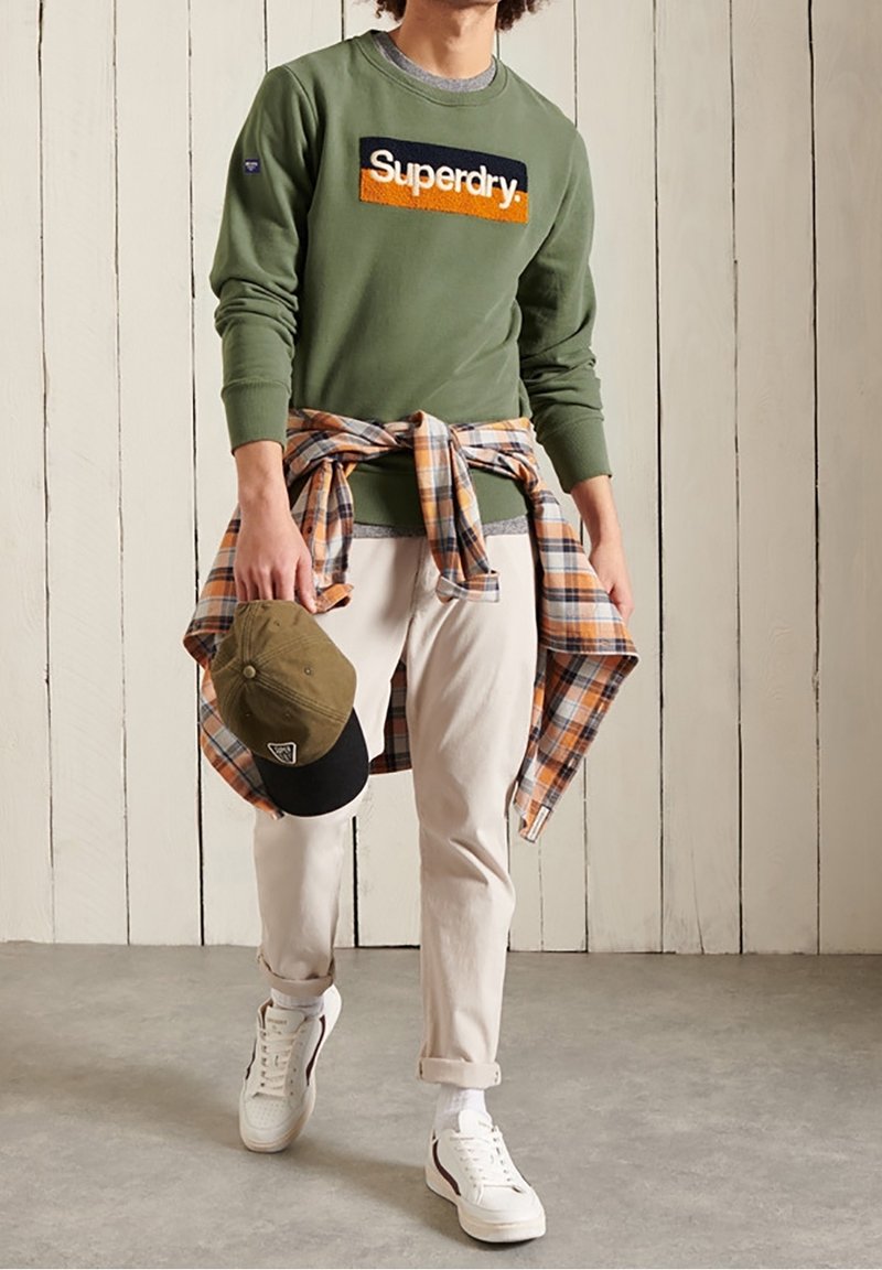 Homme portant un sweat-shirt vert Superdry, pantalon beige, baskets blanches, tenant une casquette verte et noire, avec une chemise à carreaux orange nouée autour de la taille.