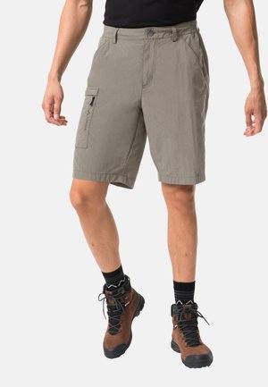 Mann trägt beige Cargoshorts, schwarzes Hemd, schwarze Socken und braune Wanderschuhe, steht vor weißem Hintergrund.