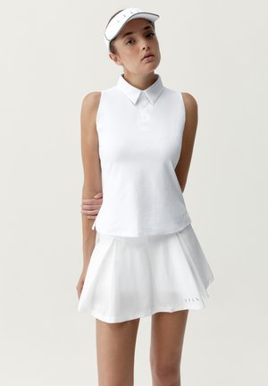 Frau trägt weißes ärmelloses Oberteil mit Kragen, weißen Faltenrock und weißen Visor, steht vor einem schlichten hellen Hintergrund.