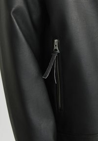 Black leather jacket sleeve showing a textured finish, featuring a silver zip closure on the side with a leather pull tab.