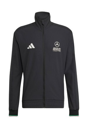 Giovane uomo con i capelli ricci che chiude una giacca nera del team AMG Petronas Formula 1 sopra una camicia bianca.
