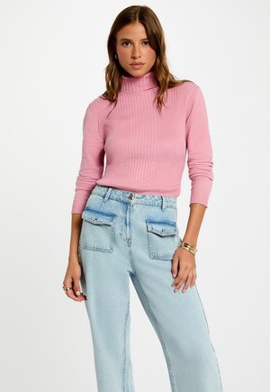 Donna che indossa un maglione a collo alto rosa a coste e jeans a vita alta azzurri con grandi tasche frontali, in posa contro uno sfondo bianco.