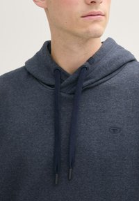 Felpa con cappuccio di un blu scuro, realizzata in tessuto morbido, con scollo incrociato e lacci navy regolabili. Dettaglio del logo discreto visibile.
