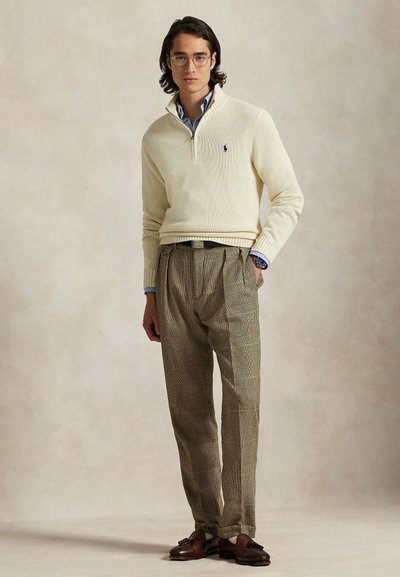 Mees, kes kannab ümarate prillidega, kreemikat kõrge kaelusega lukuga kampsunit, sinist särki, ruudulisi pükse ja pruune tuttidega mokasiine, seisab ühe käega taskus.