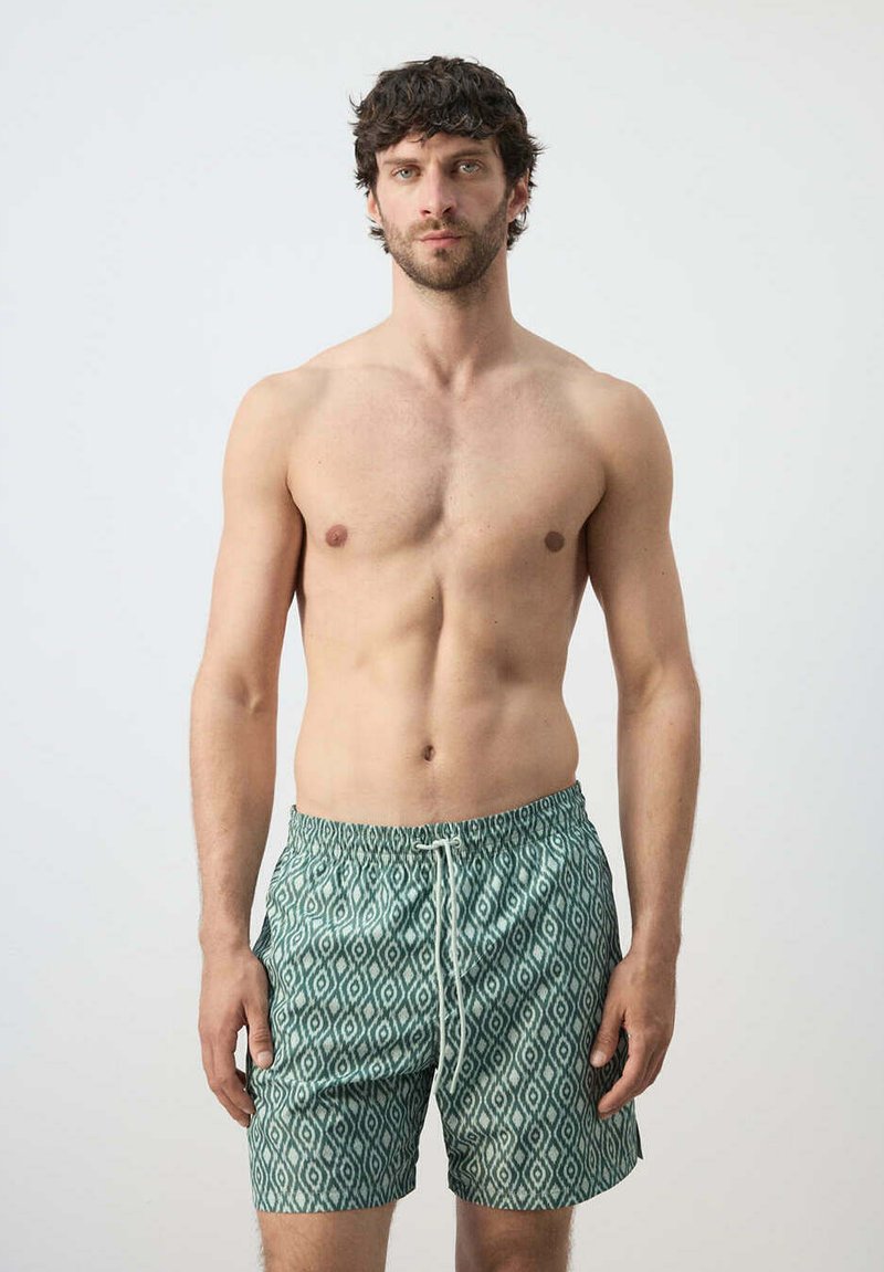 Homme aux cheveux foncés et à la barbe, portant un short de bain vert à motifs, debout devant un fond clair uni.