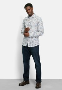 Mann trägt ein weißes, blumiges Langarmhemd, dunkelblaue Jeans und braune Slipper und steht vor einem schlichten Hintergrund.