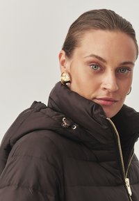 Sort pufjakke med høj krave, quiltet design og detaljer i guld på lynlåsen. Modellen bærer store guldøreringe, som fremhæver en glat tekstur.