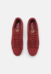 Punaiset mokkanahkakengät, joissa on teksturoitu viimeistely, valkoinen kumipohja, nauhakiinnitys edessä ja kullanväriset yksityiskohdat kielen ja logon kohdalla.