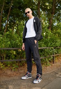 Schwarze leichte Jacke, weißes T-Shirt, schwarze Sweatpants mit Logo, graue und rote Sneakers. Außeneinstellung mit grüner Laubkulisse im Hintergrund.