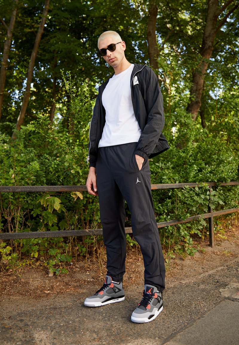 Schwarze leichte Jacke, weißes T-Shirt, schwarze Sweatpants mit Logo, graue und rote Sneakers. Außeneinstellung mit grüner Laubkulisse im Hintergrund.
