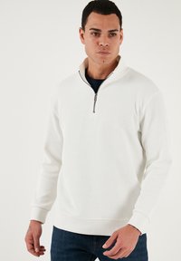 Biely rebrovaný sveter s vysokým zipsom a dlhými rukávmi, s textúrovanou tkaninou a jemne priliehavým strihom.