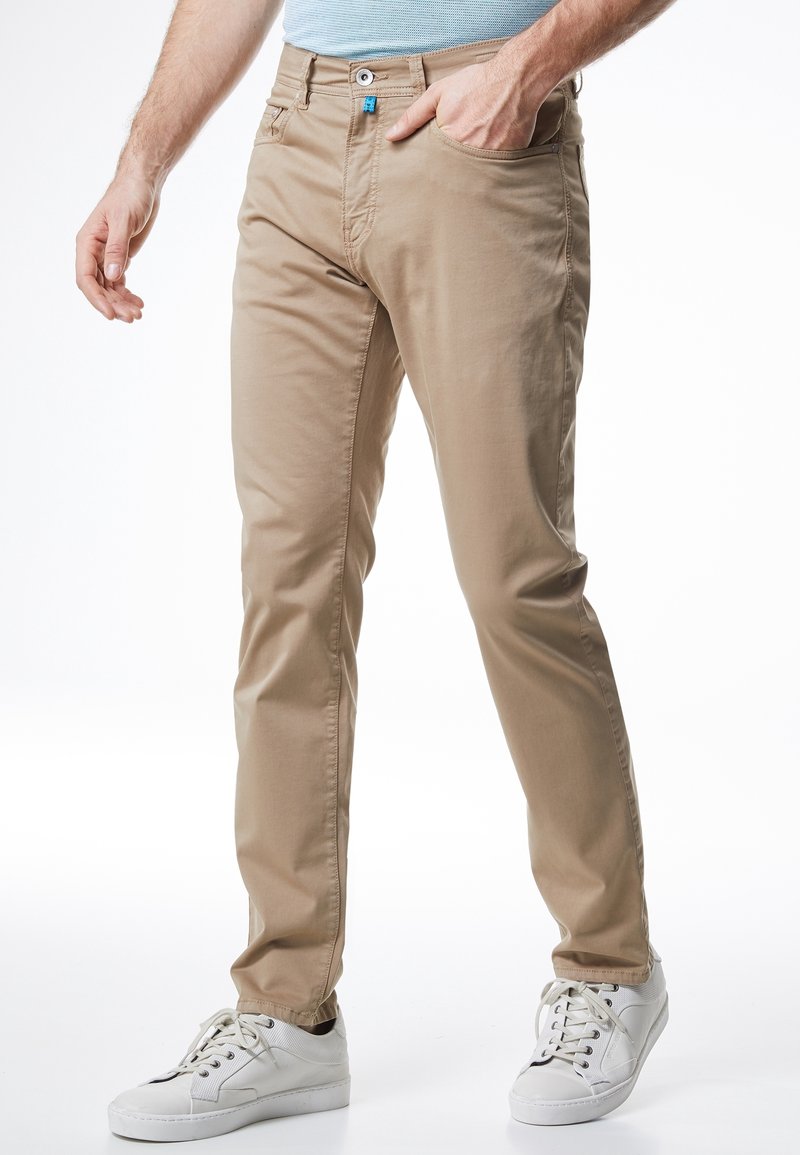Beige chinos suoran istuvuuden kanssa, joissa on tasainen etuosa, kaksi sivutaskua ja pieni sininen korostuspainike, yhdistettynä valkoisiin lenkkareihin.