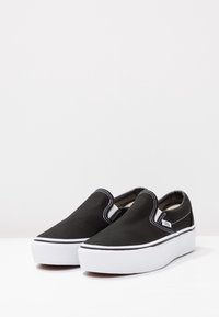 Un paio di sneaker Vans nere senza lacci con accenti elastici bianchi ai lati e spesse suole in gomma bianca su una superficie bianca.