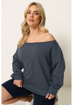 Lyshåret kvinde iført mørkegrå sweatshirt med bare skuldre, sorte shorts og hvide sneakers, poserer mod en ensfarvet lys baggrund.