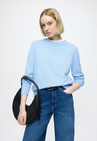 Jasnoniebieski sweter z okrągłym dekoltem, zestawiony z niebieskimi dżinsami z wysokim stanem. W dłoni trzyma czarną, zakrzywioną torebkę z długim paskiem.