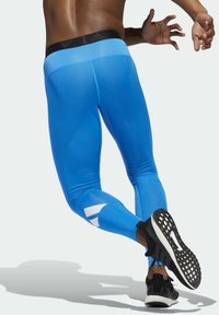Niebieskie legginsy sportowe wykonane z elastycznego materiału, z czarną talią i białymi akcentami po bokach. Noszone z czarnymi butami biegowymi.