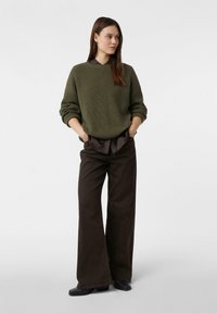 Olivgrüner, gerippter Pullover mit überschnittenen Schultern, kombiniert mit dunkelbraunen, weit geschnittenen Hosen. Das Modell steht mit den Händen in den Taschen.