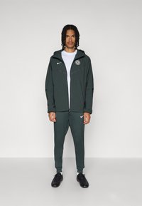 Dark green Nike tracksuit featuring a zip-up jacket with hood, white logo, and matching tapered pants, worn with black athletic shoes.