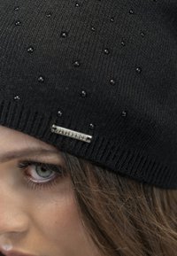 Zwarte gebreide beanie met kleine ronde versieringen en een metalen logo tag. De textuur is geribbeld en het ontwerp is aansluitend rond het hoofd.