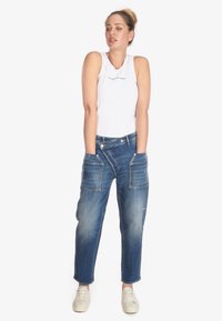Camiseta sin mangas blanca con textura acanalada y texto gráfico, combinada con jeans de mezclilla azul de corte relajado que tienen bolsillos frontales y un ligero desvanecimiento. Se llevan zapatillas neutras.
