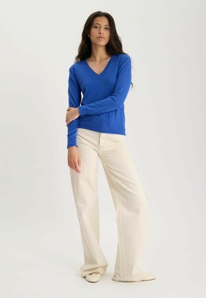 Femme portant un pull bleu col en V, un pantalon crème à larges jambes et des chaussures crème, debout avec un bras croisé sur son corps.