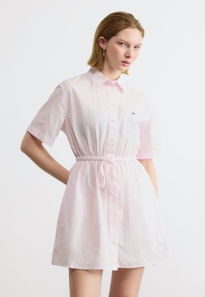 Fiatal nő rövid ujjú, világos rózsaszín gombos ruhában, derékban megkötve, fehér háttér előtt áll, kezét a csípőjén tartva.