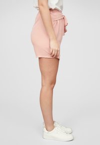 Shorts roses avec une finition texturée, dotés d'une taille nouée, de poches latérales et d'une silhouette ajustée, associés à des baskets blanches.