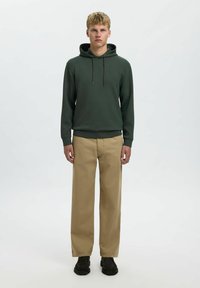 Felpa verde con cappuccio e cordoncino, polsini e fondo a coste, abbinata a pantaloni beige a gamba larga e scarpe nere, su uno sfondo neutro.