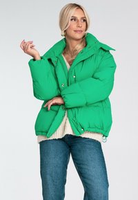 Zielona kurtka puchowa z dużym kołnierzem, zatrzaskami i elastycznym dołem. Noszona na beżowym swetrze, w połączeniu z niebieskimi dżinsami.