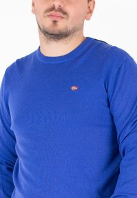 Niebieski sweter z długim rękawem, dziany, z okrągłym dekoltem i małym emblematem norweskiej flagi na klatce piersiowej. Miękka faktura, dopasowany krój.