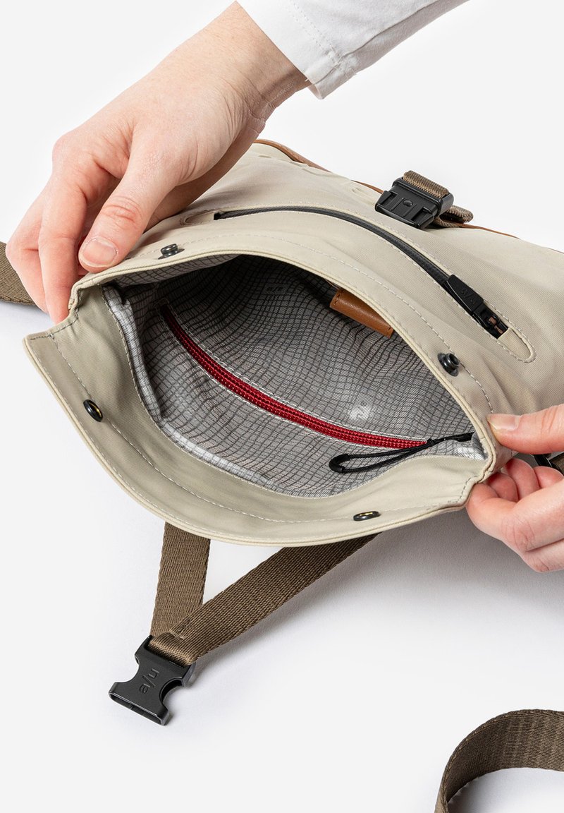 Interieur van een beige tas met een grijs patroon aan de voering, rood ritsvak en bruin hengsel. Bovenste ritssluiting en zwarte hardware details zichtbaar.