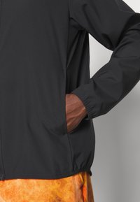 Hand in pocket of black jacket with elastic cuff, paired with orange tie-dye pants against a light gray background.