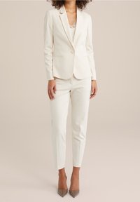 Kremowy garnitur skrojony na miarę, z dopasowaną marynarką i slim fit spodniami, wykonany z gładkiego materiału. Posiada zapięcie na jeden guzik i minimalistyczny design.