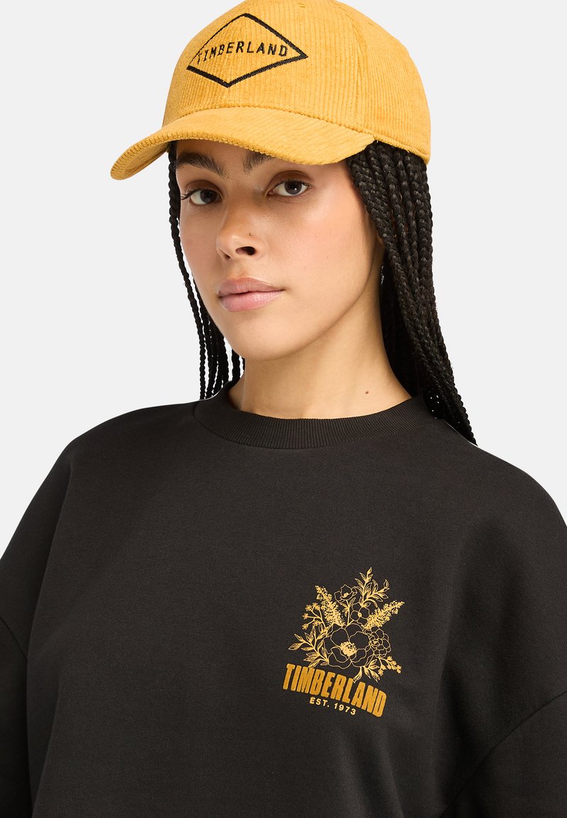 Cappello in corduroy giallo con logo nero; felpa nera con ricami floreali gialli e testo "TIMBERLAND EST. 1973". Collo ampio, vestibilità rilassata.