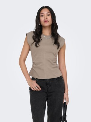 Vrouw met lang donker haar, gekleed in een mouwloos taupekleurig topje en zwarte jeans, met een zwarte handtas tegen een effen lichte achtergrond.