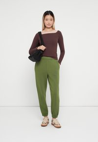 Top marrón de manga larga con canalé y escote cuadrado, combinado con pantalones de chándal verdes. Un bolso negro de hombro y zapatillas deportivas con estampado completan el look.