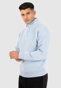Sweatshirt bleu clair à col zippé, avec des poignets et un ourlet côtelés. Fabriqué en tissu doux avec une texture lisse.