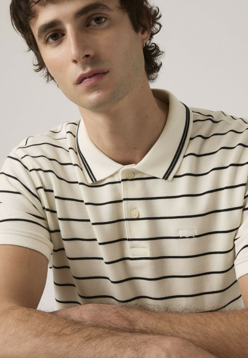 Joven con cabello corto y rizado, que lleva una camisa tipo polo crema con rayas horizontales negras, apoyando los brazos cruzados sobre una superficie plana.