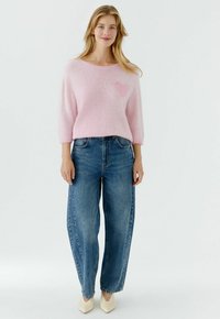 Femme debout, les mains le long du corps, portant un pull duveteux rose clair avec un détail en forme de cœur, un jean bleu à jambes larges et des chaussures pointues crème.