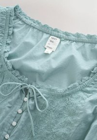 Blouse bleu clair bordée de dentelle avec de petits boutons blancs et un nœud à l’encolure, montrant une étiquette blanche portant l’inscription « NEXT time to dream ».