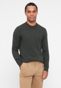 Un pull tricoté de couleur vert foncé avec un col rond et des poignets côtelés, associé à un pantalon beige clair. La texture semble douce et confortable.