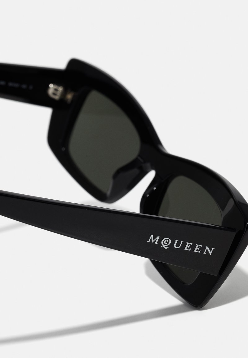 Occhiali da sole neri con montatura angolare, lenti scure e logo "MQUEEN" sul braccio. Finitura lucida e dettagli metallici alla cerniera.